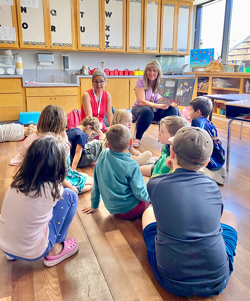 About-Teach-1 Teacher and Assistance reading a book to Kindergarteners